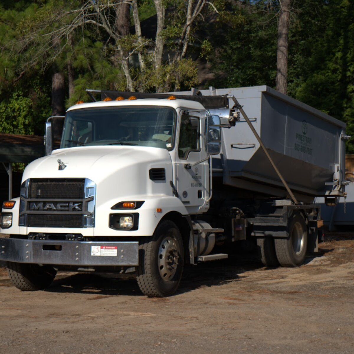 White Mack dump truck with a loaded dump bed parked on a dirt lot, trees in the background and a utility trailer hitching a load behind the cab