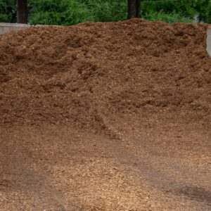 Large mound of brown wood mulch piled outdoors with green trees in the background