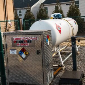 Propane storage area with a large white tank and a diamond-shaped hazard label, next to a metal utility cabinet labeled 'Flammable Gas Propane No Smoking' and warning symbols.