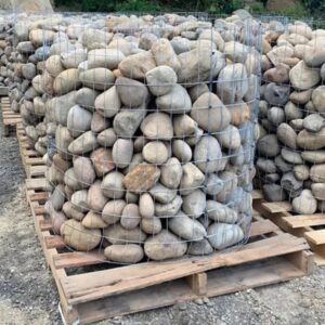 Large round river rocks packed inside a cylindrical wire-gabion basket on a wooden pallet in a gravel lot.