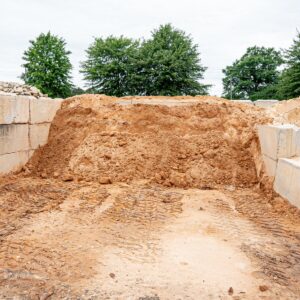 Excavation pit with piled dirt and concrete block walls at a construction site, muddy ground with tire tracks in foreground.